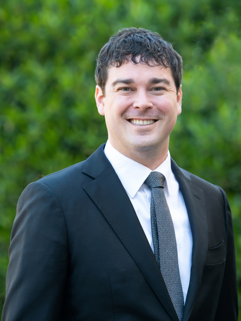 Brett Vaughn, Attorney and Partner at Nigh Goldenberg Raso & Vaughn, PLLC, smiling in front of a garden backdrop.